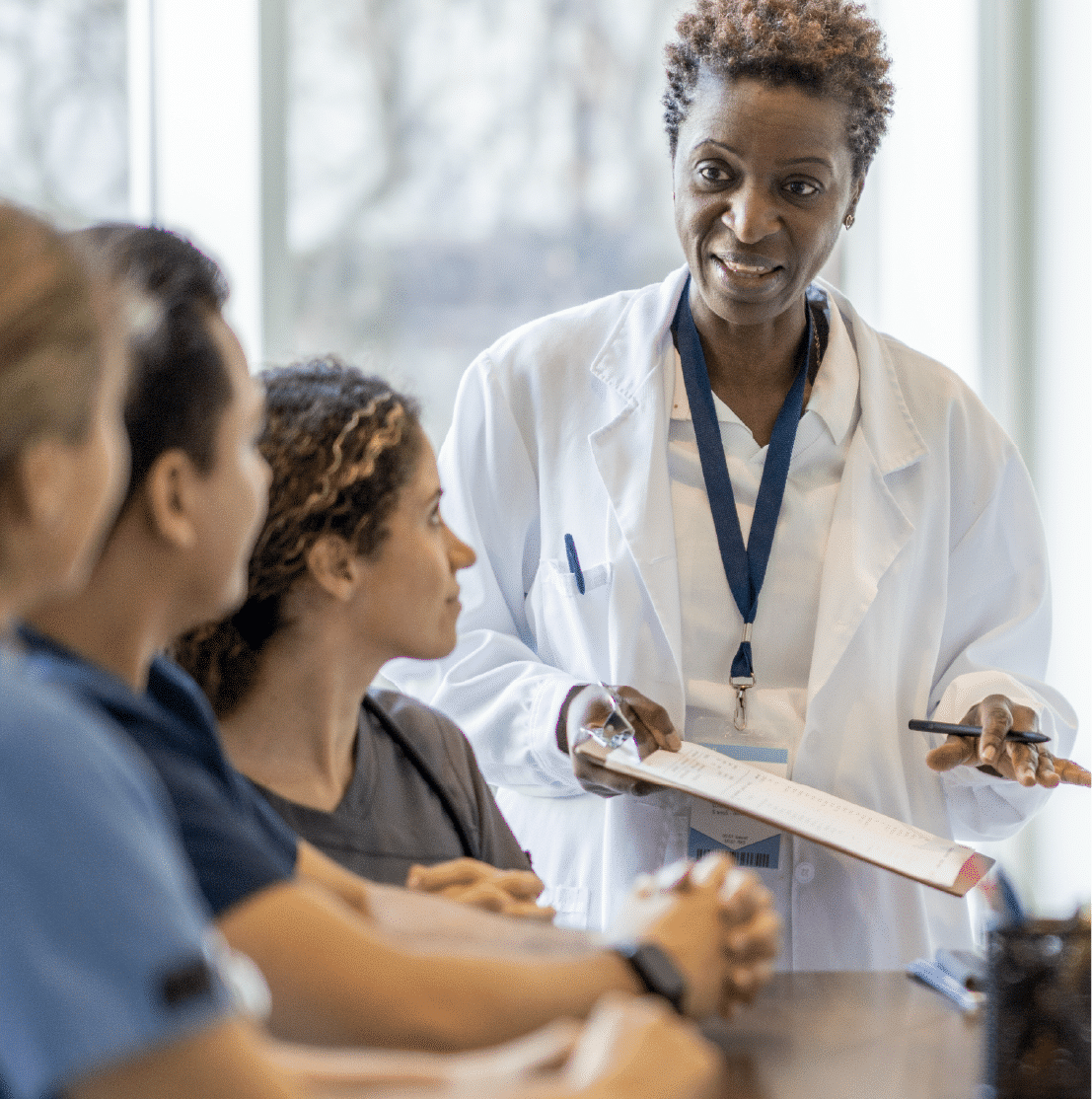 A group of medical people in a meeting. A woman in a white lab coat is standing and talking to three staff.