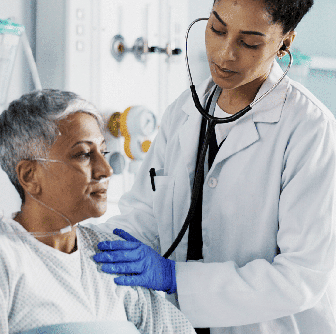 A doctor listening to a woman's heart. The woman is wearing a hospital gown and has an oxygen tube in her nose.